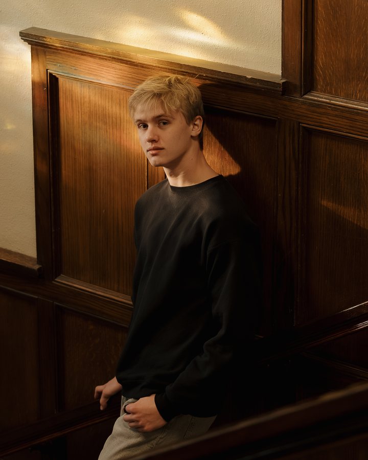 Joel Haley leaning on a wood-paneled staircase in warm window light